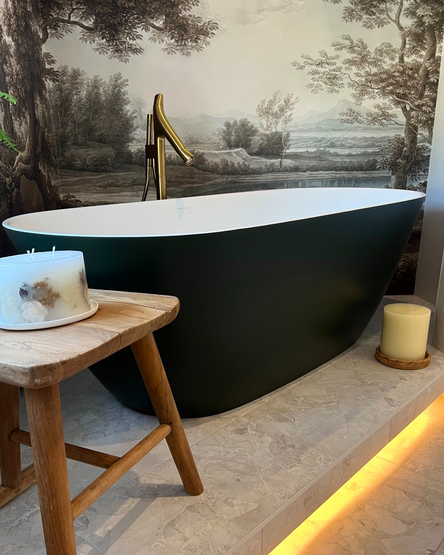 Dark freestanding bath with a brass tap in front of a landscape mural, displayed in the showroom